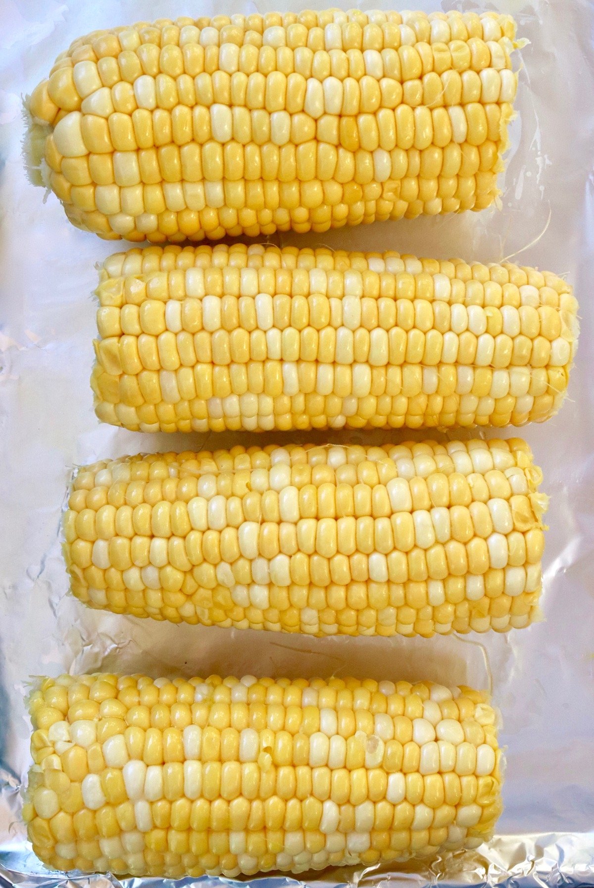 Four ears of raw corn on foil-lined baking sheet.