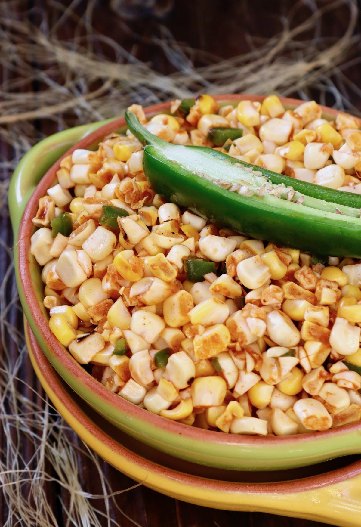 Green bowl filled with fire roasted corn and half of a raw jalapeno surrounded by corn silk.