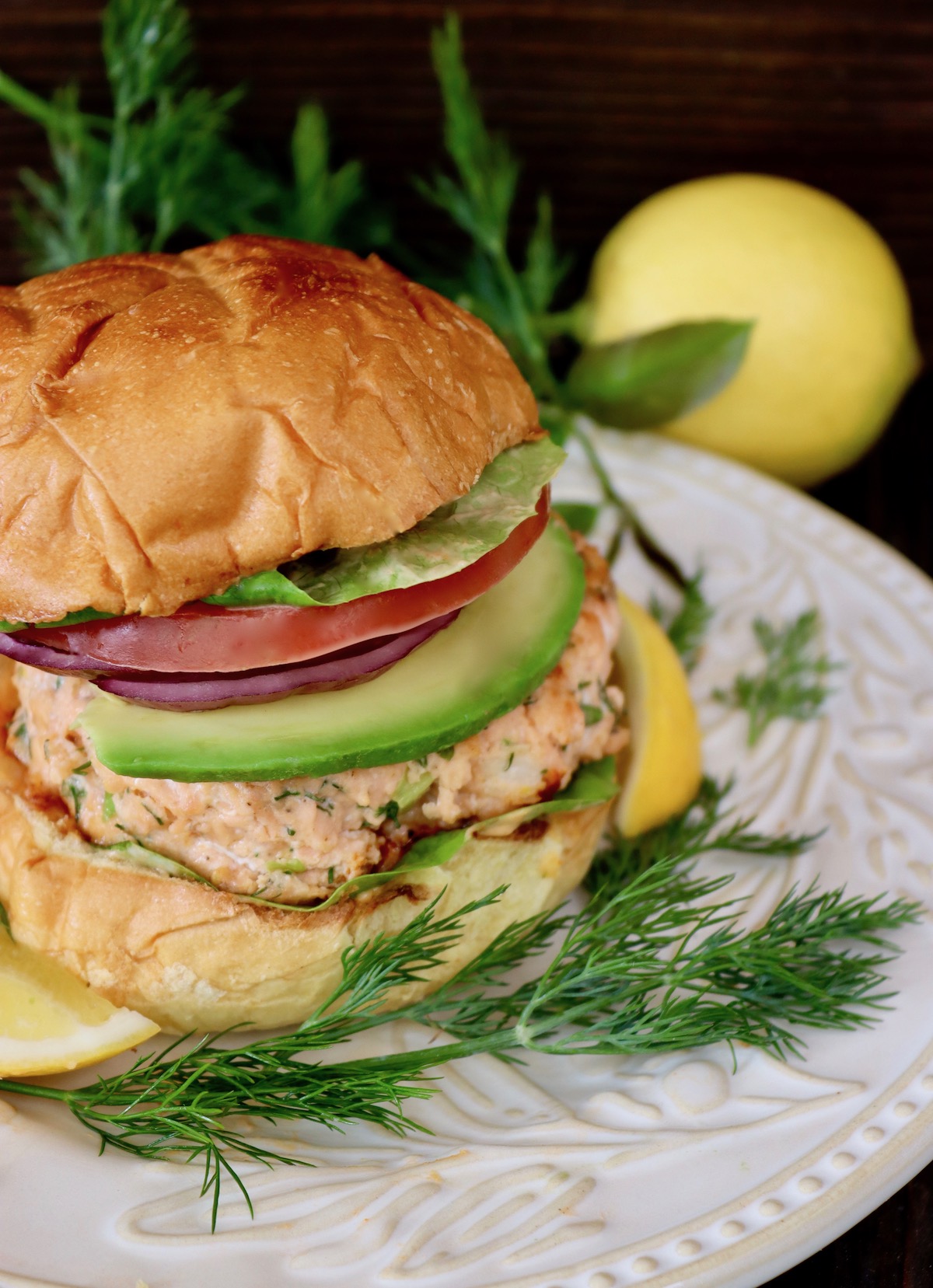 Salmon burger with Brioche bun with tomato, onion and avocado on white plate.