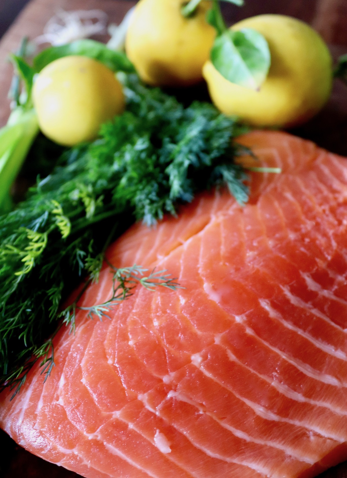 Close up of large raw salmon fillet with fresh dill and 3 whole lemons on dark wood surface.