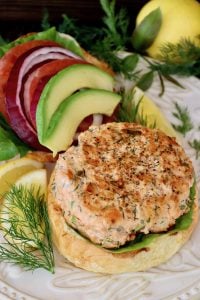 Open-salmon burger with slices of avocado tomato, red onion, dill and lemons on white plate.