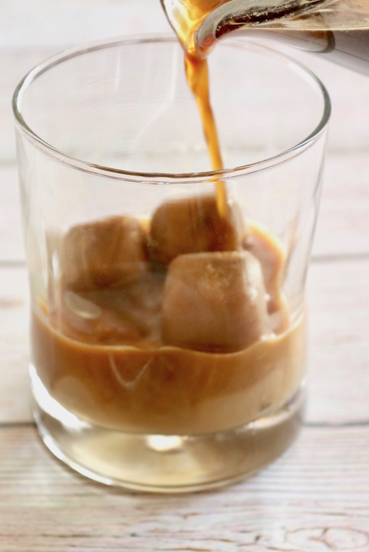 Clear drinking glass with creamy coffee ice cubes and coffee being poured into it.