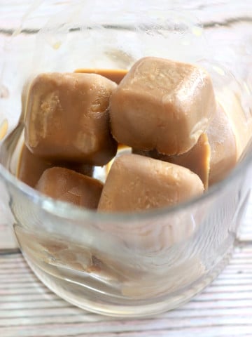 Clear drinking glass with several creamy coffee ice cubes in it.