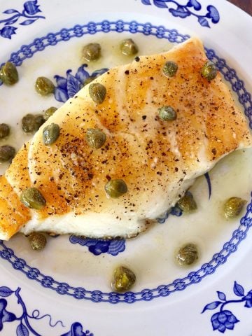 Top view of one Chilean Sea Bass fillet with a caper butter sauce on top, on a what and blue plate.