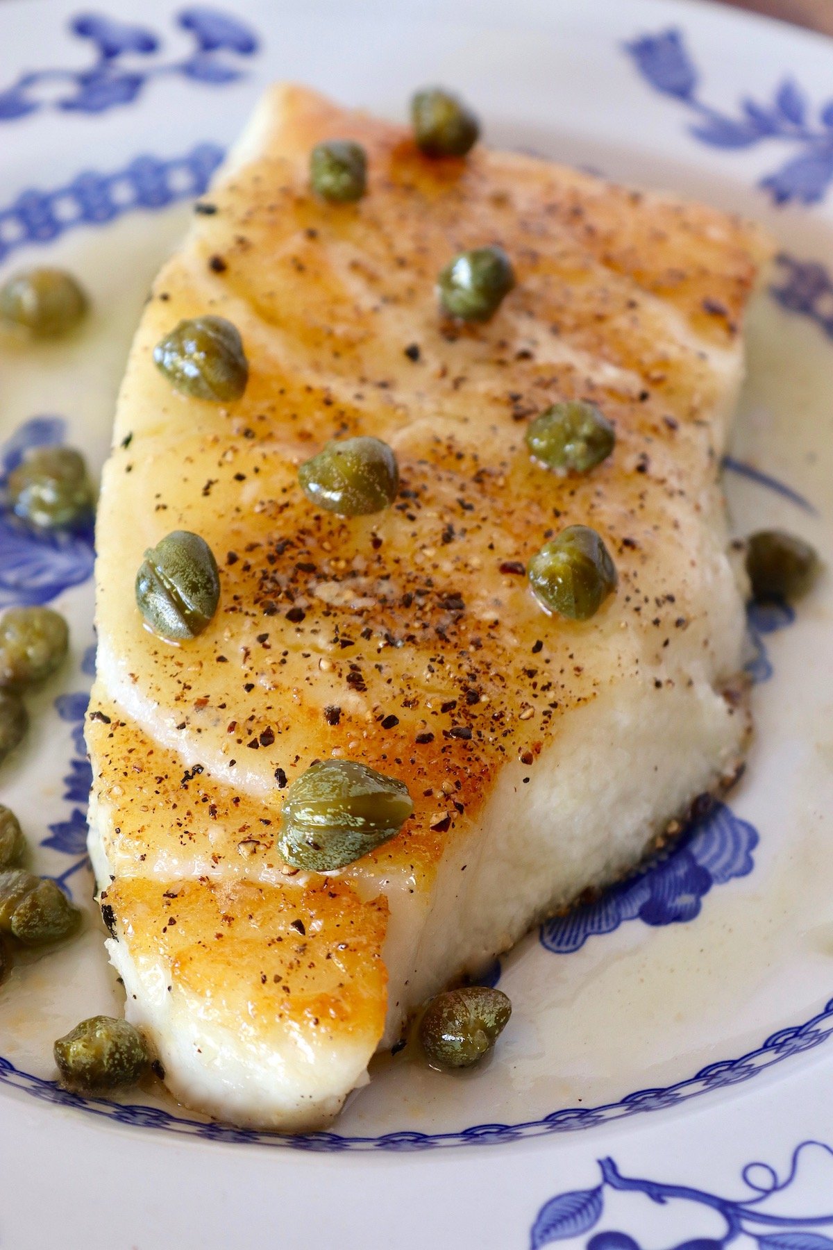 One fillet of golden pan-seared sea bass with capers on top, on blue and white china.