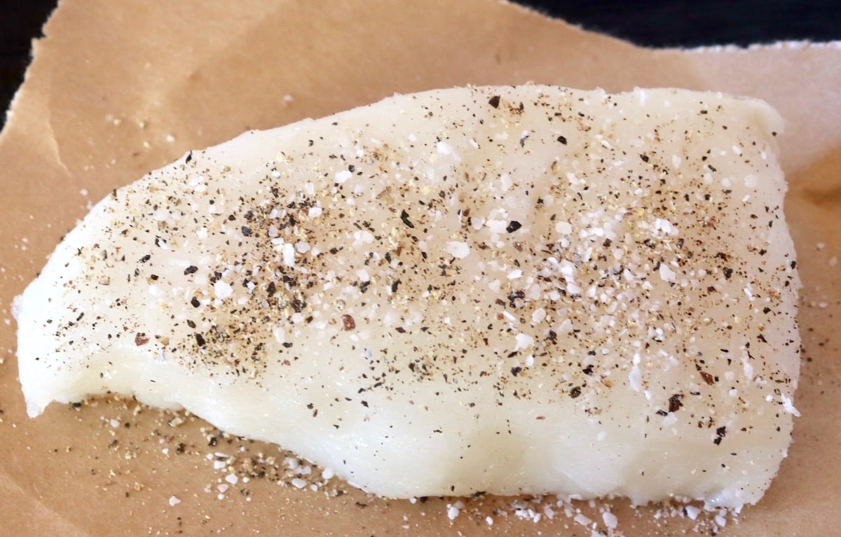 Raw fillet of sea bass sprinkled with salt and pepper on brown wax paper.