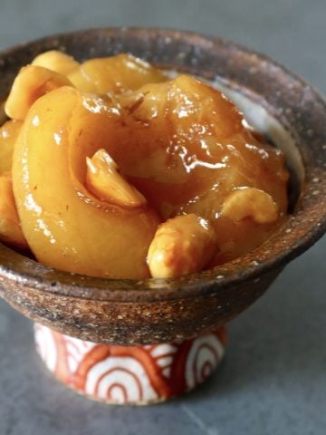 Small brown ceramic bowl with orange-patterned stem, filled with baked caramel apples and hazelnuts.