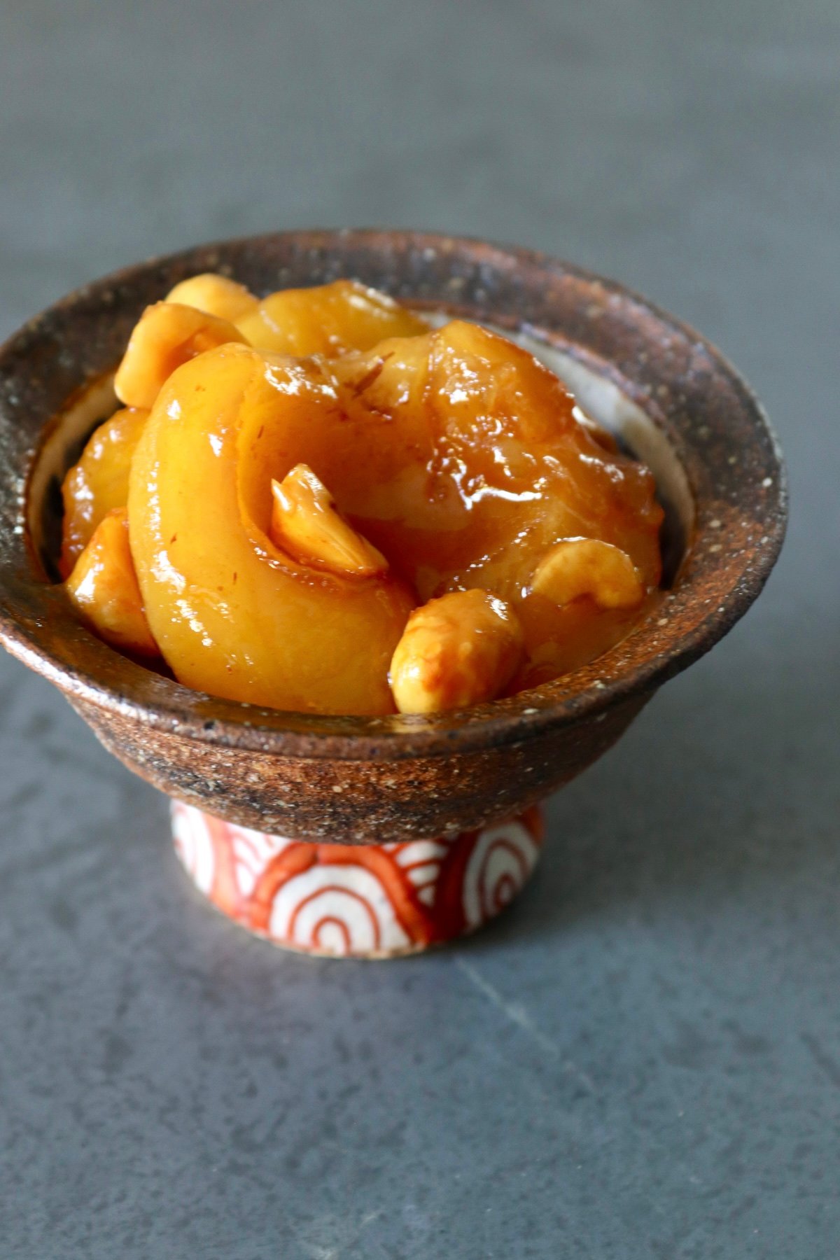 Small brown ceramic bowl with orange-patterned stem, filled with baked caramel apples and hazelnuts.