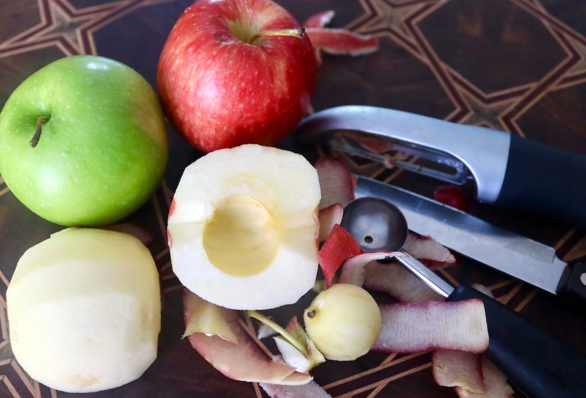 One apple that's peeled and cored with peels around it and one red, and one green apple.