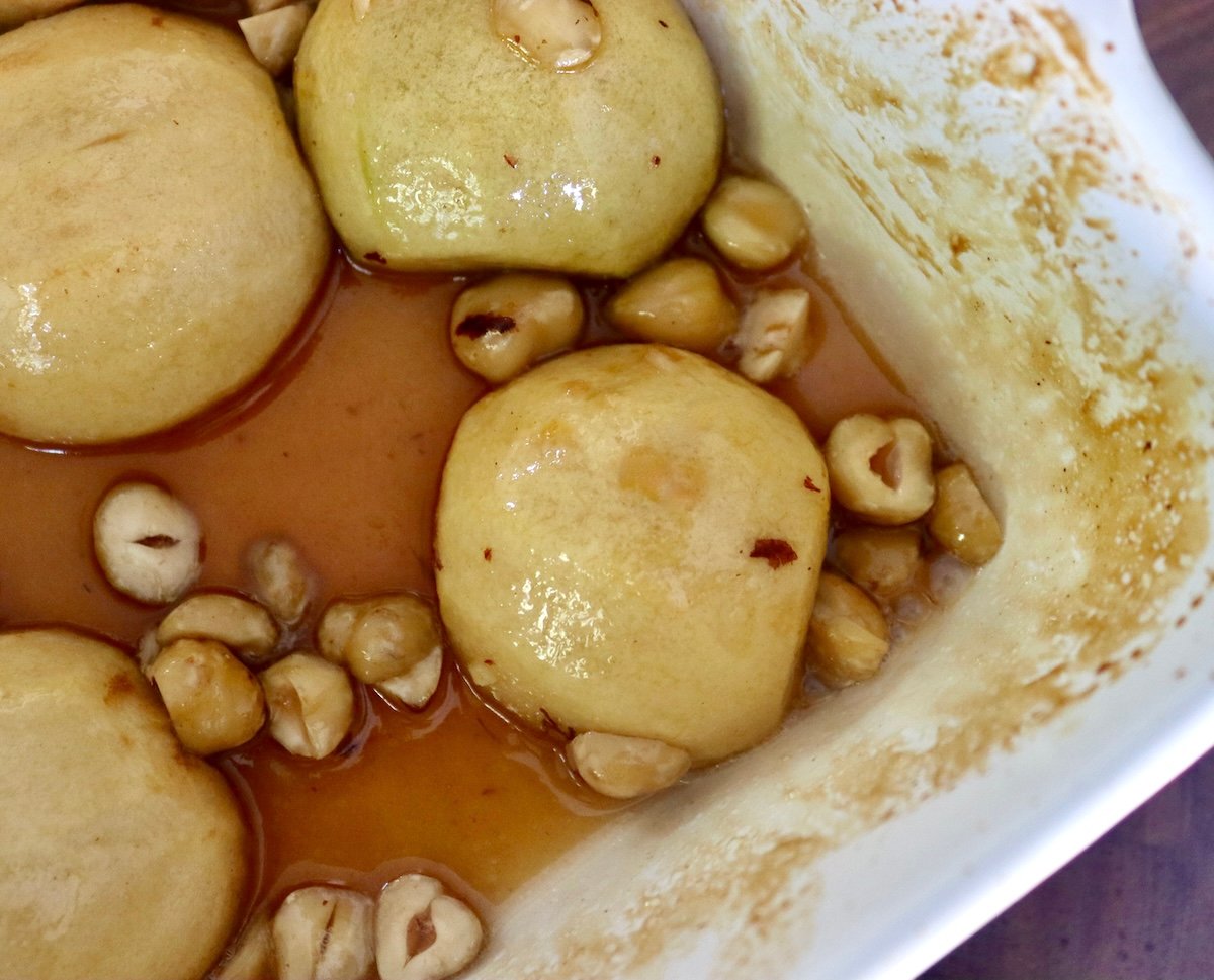 Peeled half-baked apple halves, round sides up, in a baking dish with syrup and hazelnuts.