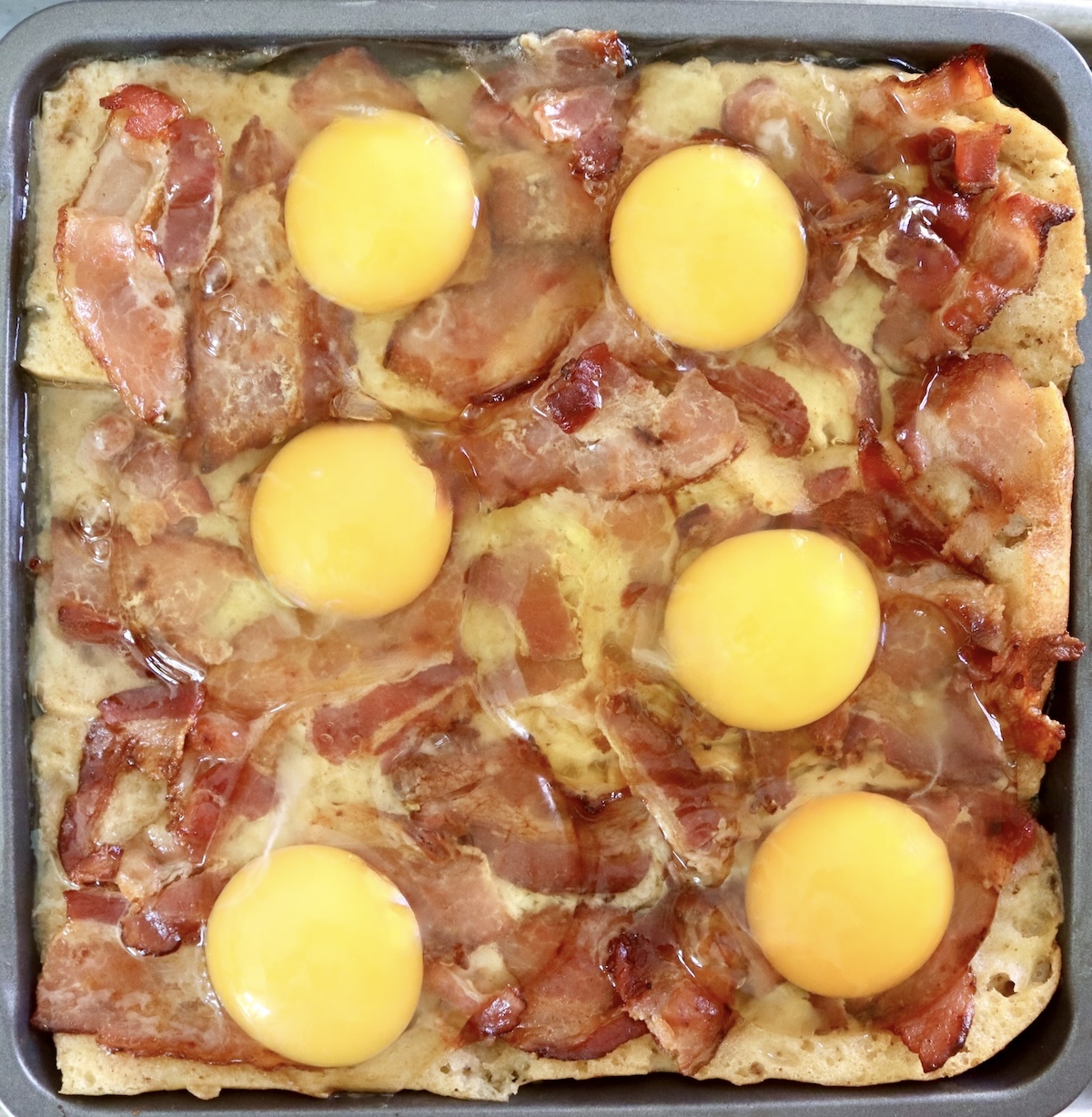 Bread with bacon pieces and raw eggs on top in square pan for French Toast Bake.