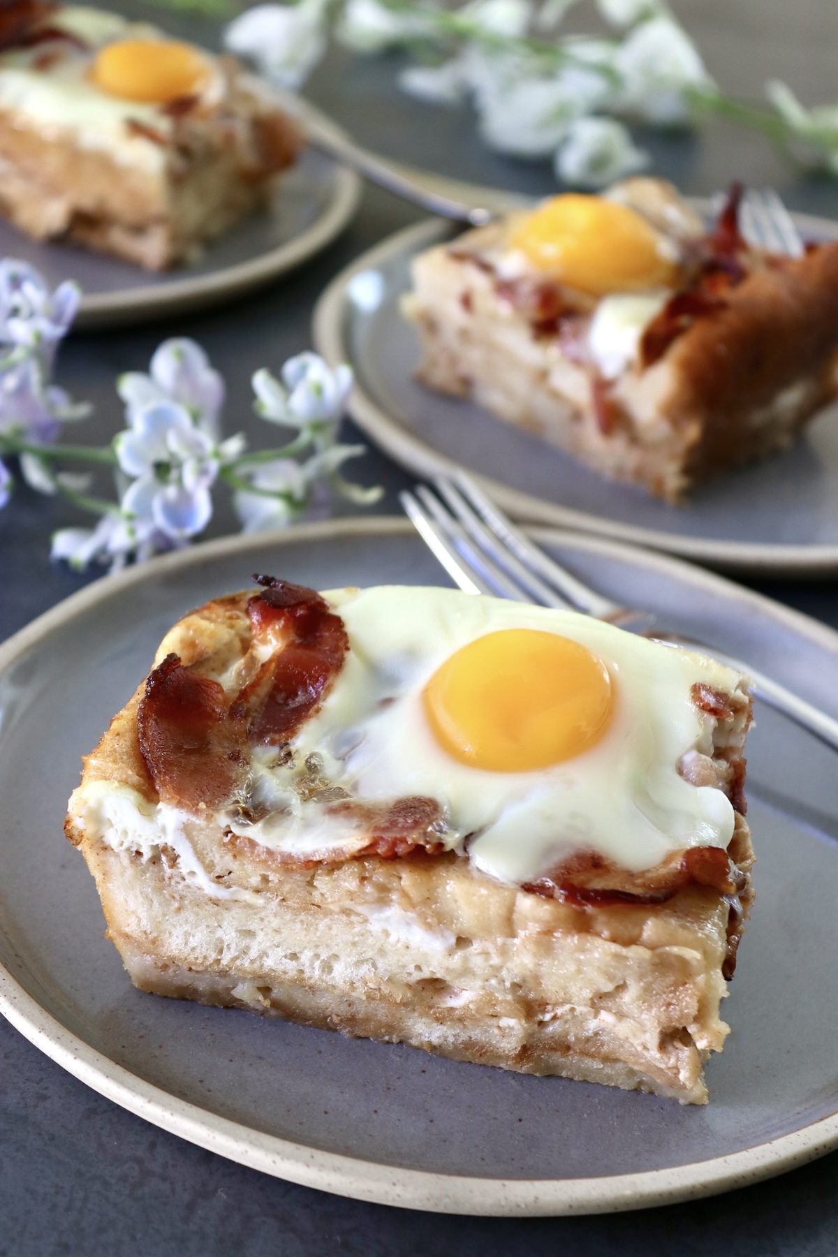 One square slice of French Toast Bake with bacon and eggs on top, on a gray plate, with two of the same behind it that are out of focus.