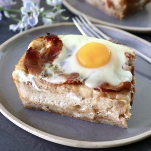 One square slice of French Toast Bake with bacon and eggs on top, on a gray plate.
