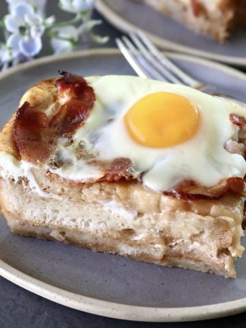One square slice of French Toast Bake with bacon and eggs on top, on a gray plate.