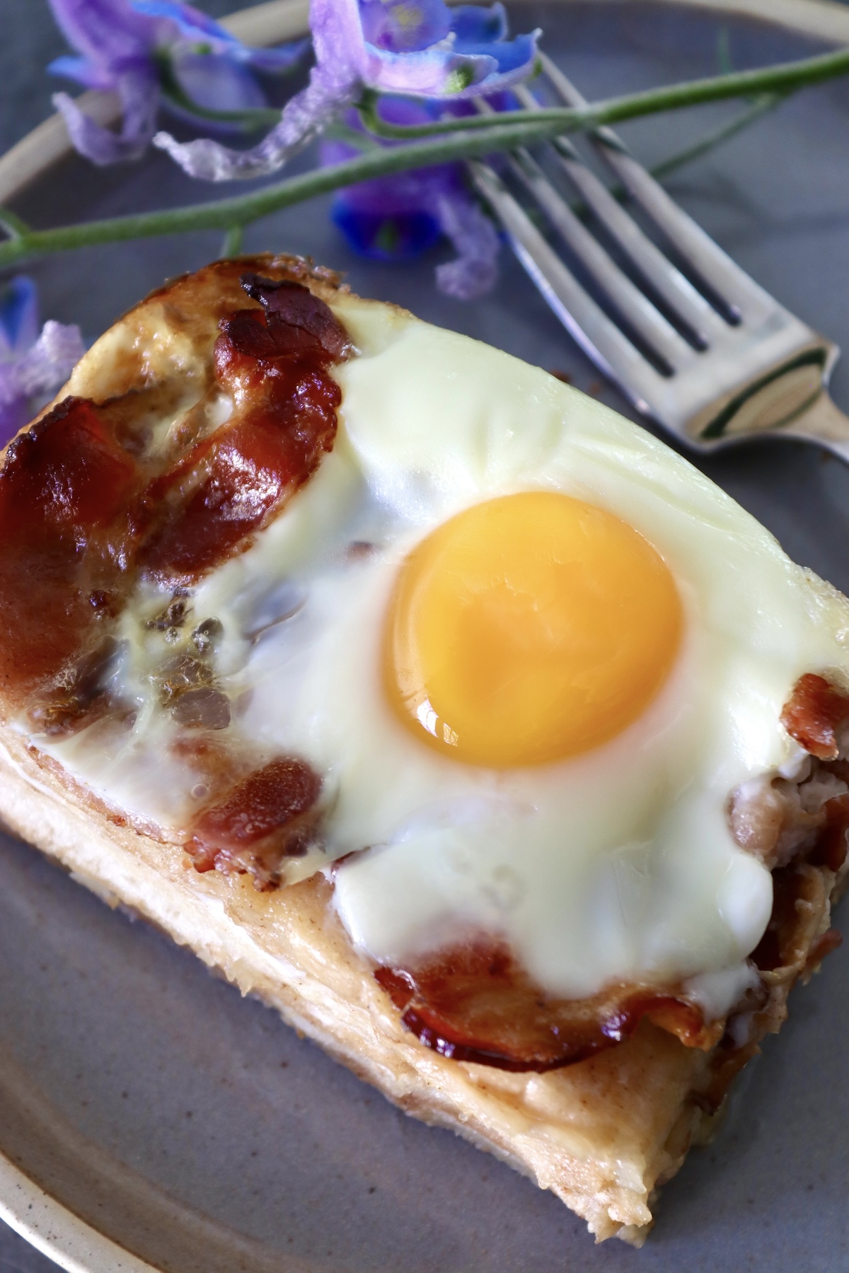Close up of one square slice of French Toast Bake with bacon and eggs on top, on a gray plate.