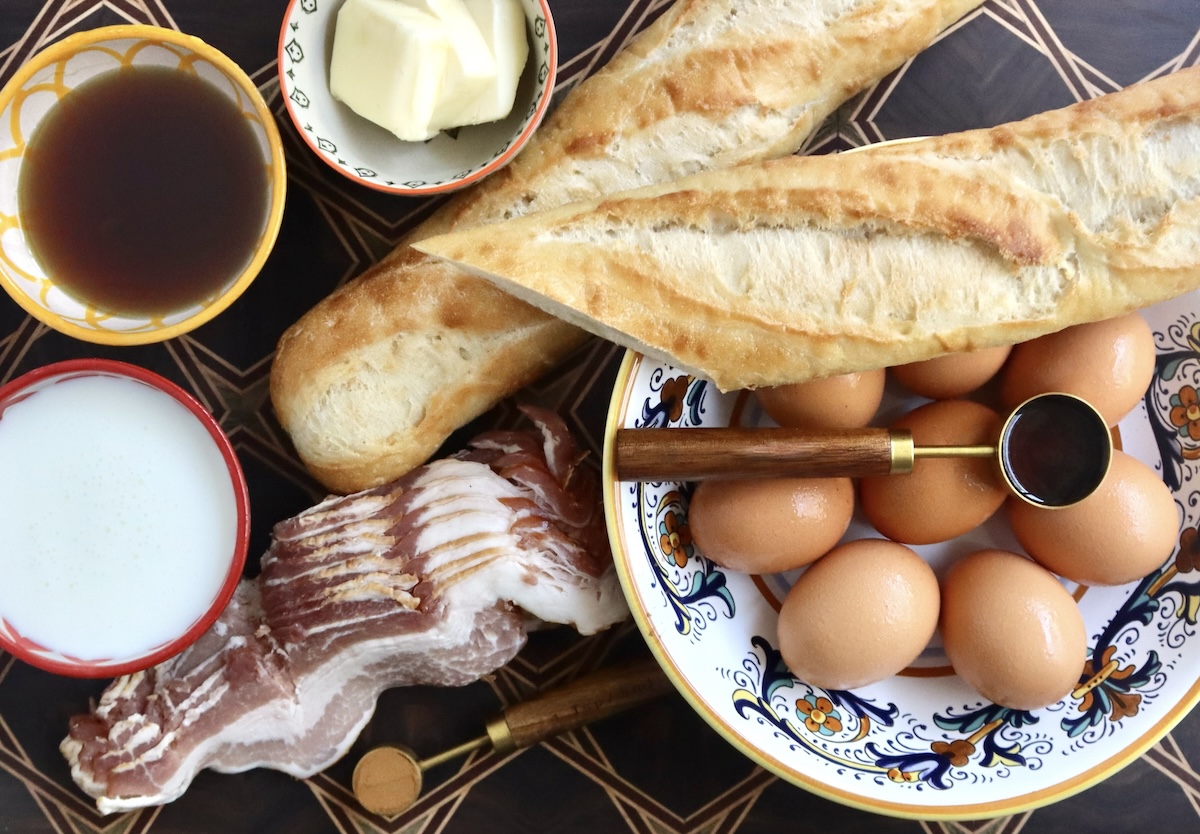 Ingredients for french toast bake with bacon and eggs, including butter, bacon, eggs, baguette, cinnamon, syrup, buttermilk and vanilla -- all on a cutting board and in bowls.