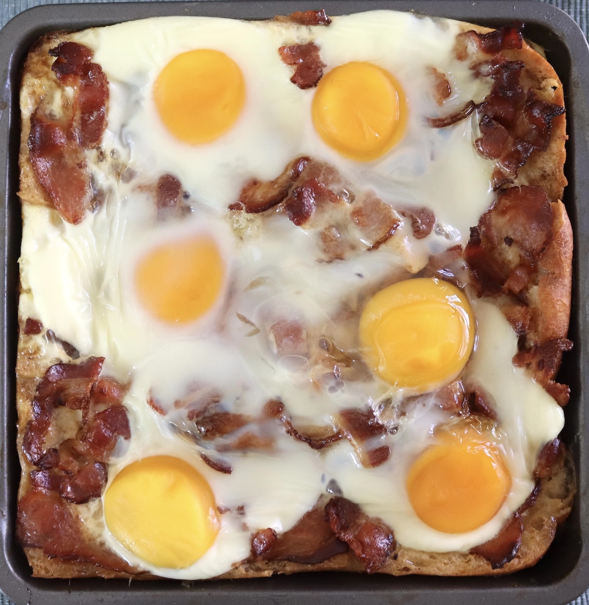 Bread with bacon pieces and cooked eggs on top in square pan for French Toast Bake.