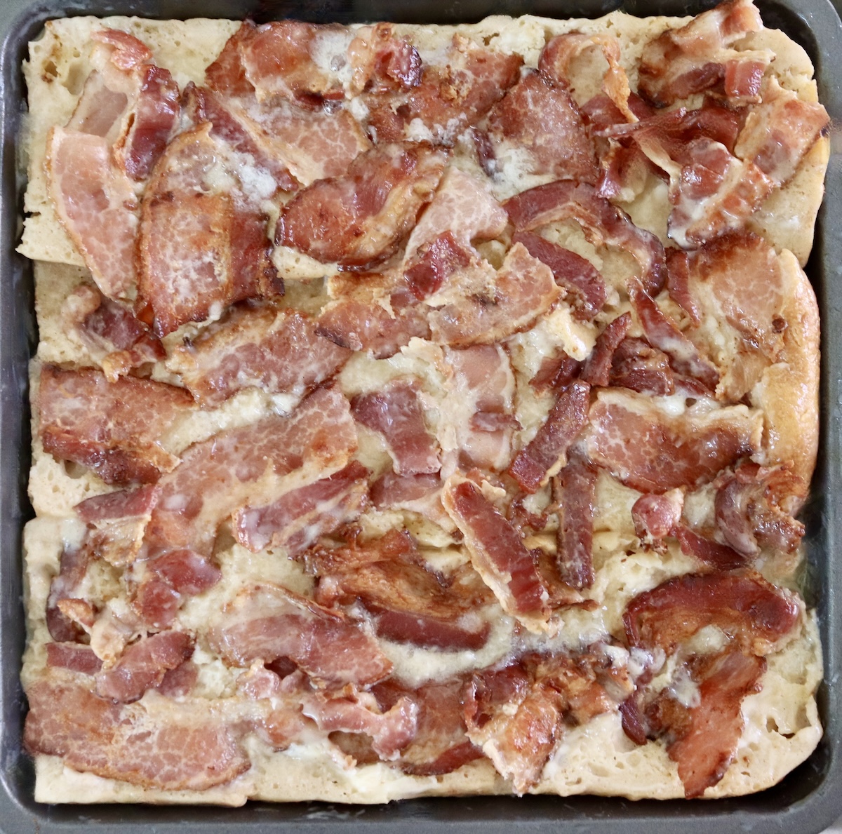 Bread with bacon pieces on top in square pan soaking for French Toast Bake.