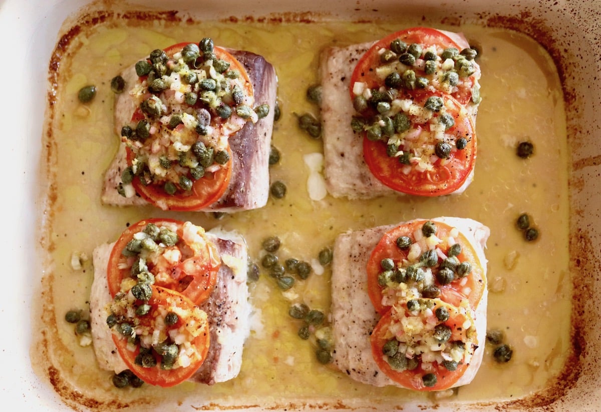 4 Baked Mahi Mahi fillets in a baking pan with oil and capers and tomato slices with caper sauce on top.