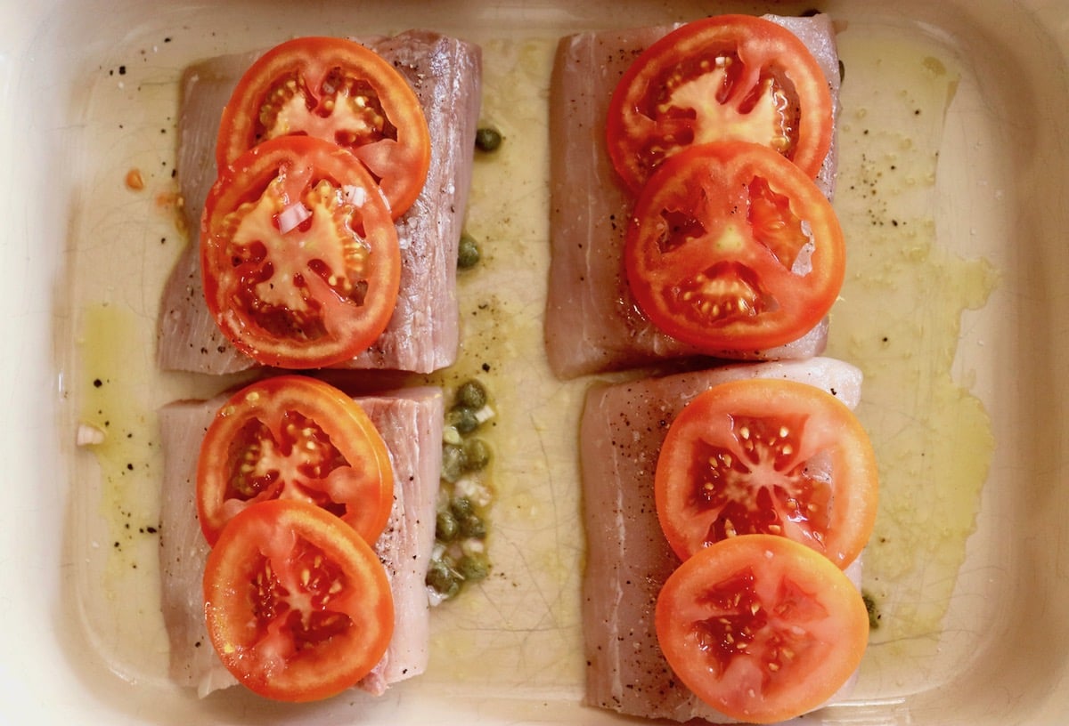 4 Mahi Mahi fillets in a baking pan with oil and capers and tomato slices on top.