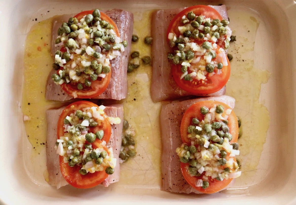 4 Mahi Mahi fillets in a baking pan with oil and capers and tomato slices on top and caper sauce on top.