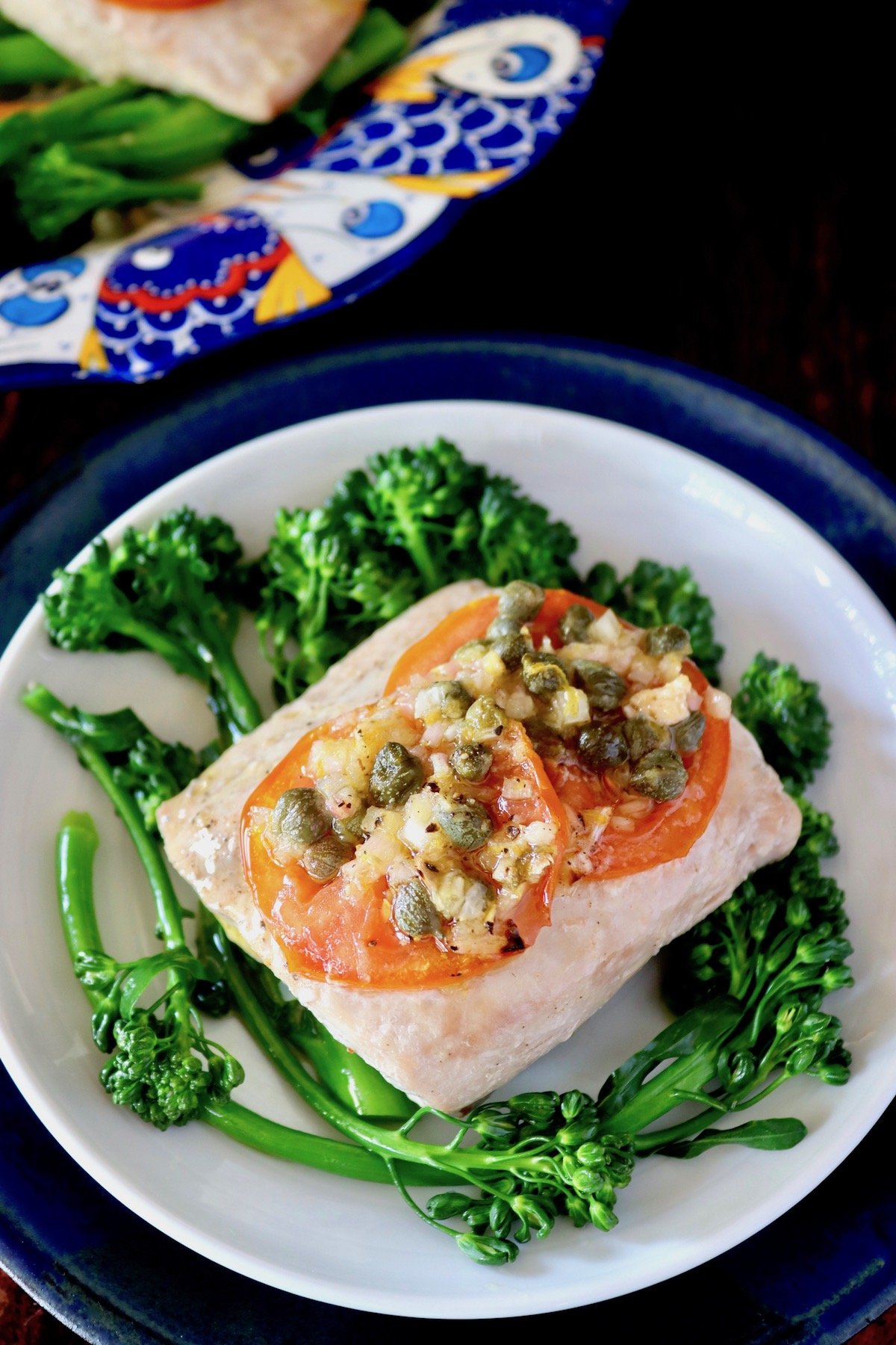 White plate with bright green Broccolini with a Mahi Mahi fillet with tomato slices on top of it.