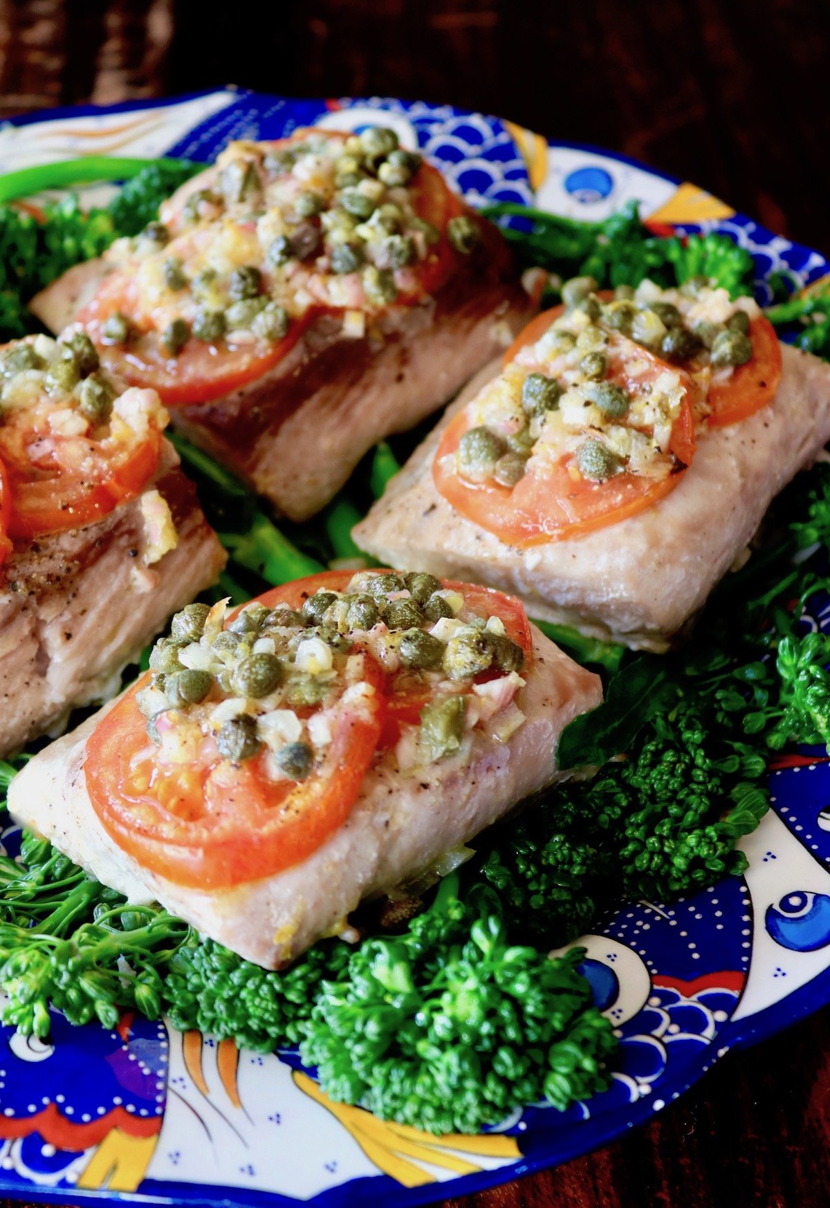 4 Baked Mahi Mahi fillets on a bright blue-rimmed plate with tomatoes and caper sauce on top.