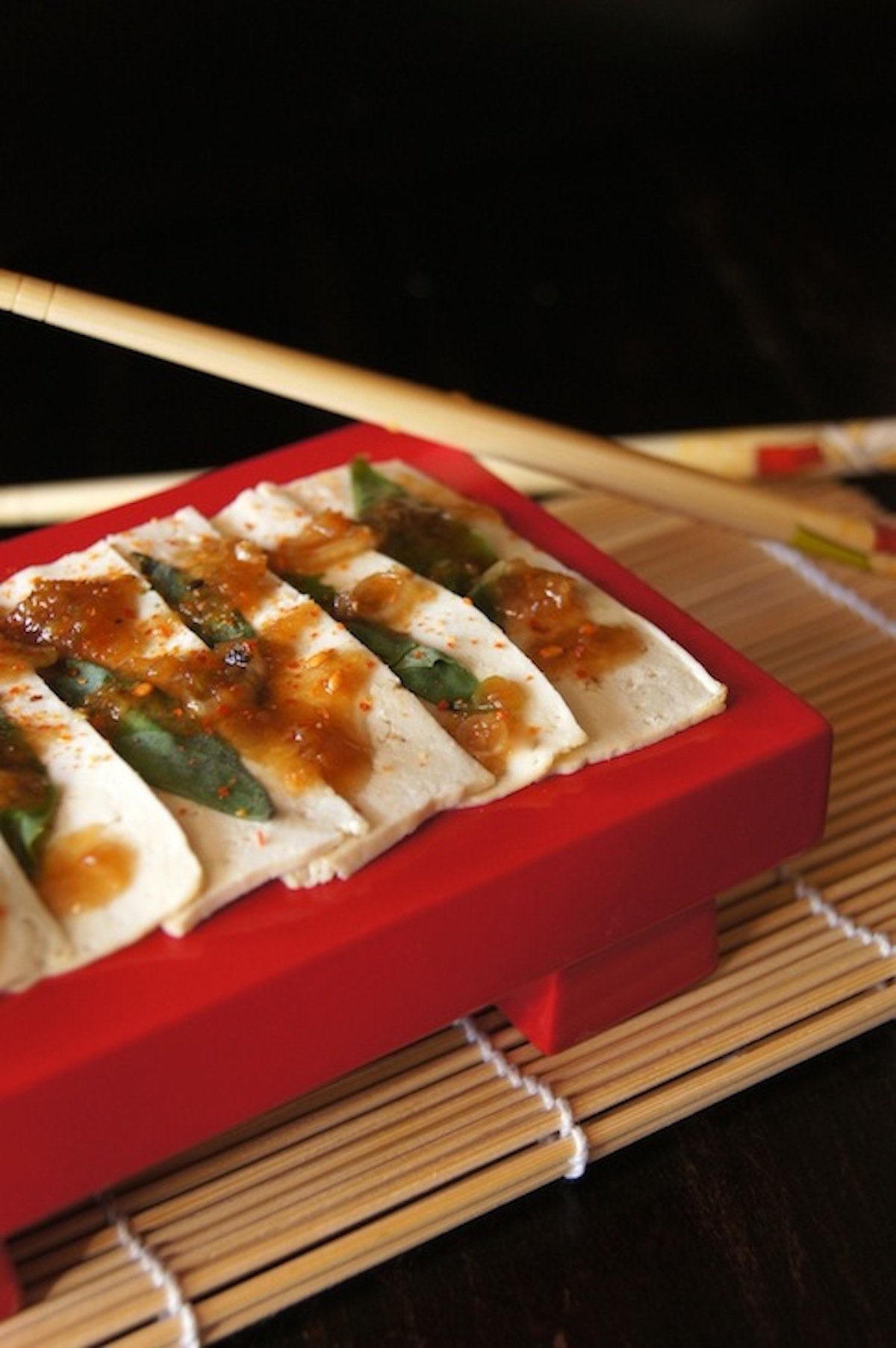 Red retangular plate with very thin slices of tofu withbasil leaves and drizzled sauce.