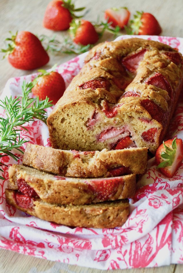 Sliced Strawbery-Rosemary Cake on a white and pink cloth with fresh rosemary sprigs and whole strawberries in background.