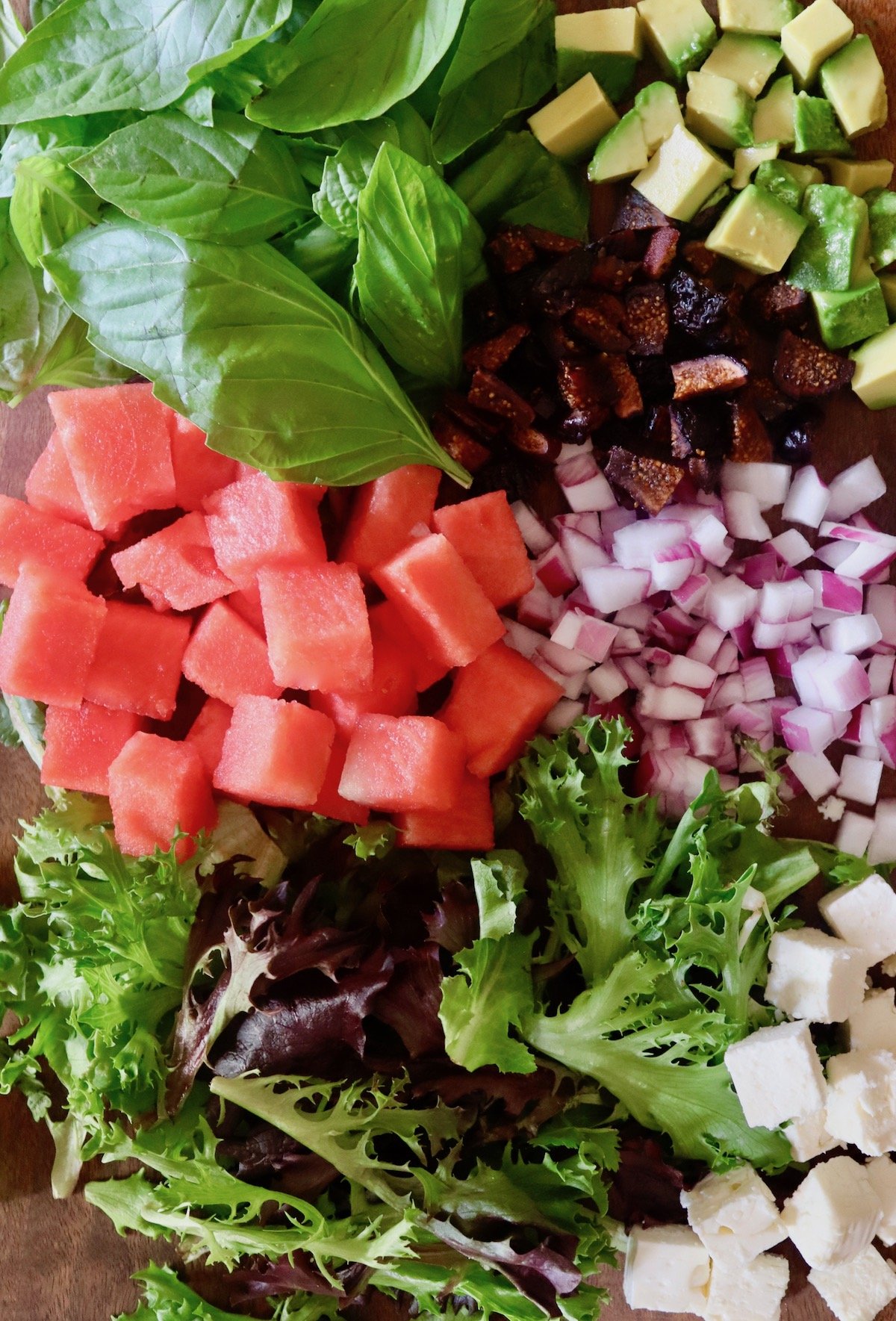 Chopped watermelon, feta, red onion, dried figs and lettuce and basil leaves on a cutting baord.