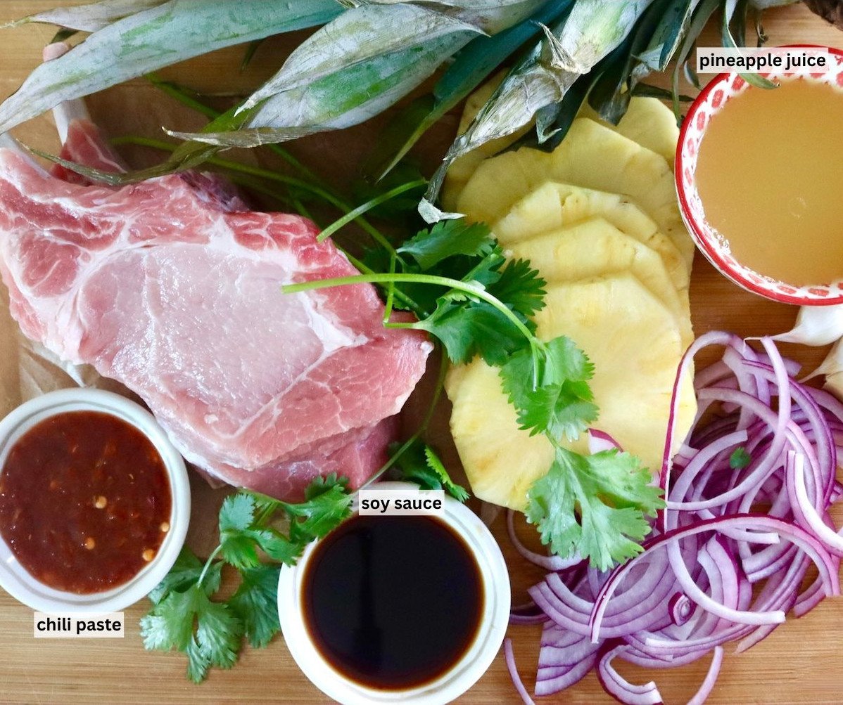 Ingredients for pineapple pork including, raw pork chops, red onion slices, pineapple juice and slices, chili paste, soy sauce and garlic -- all on a cutting board.