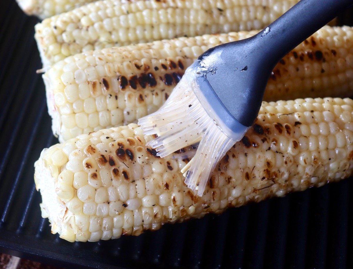 Grilled white corn being brushed with lemon pepper butter.