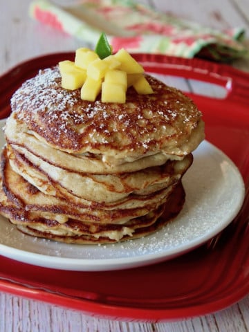 Tall stack of coconut flour pancakes on a whitle place iwth mango on top