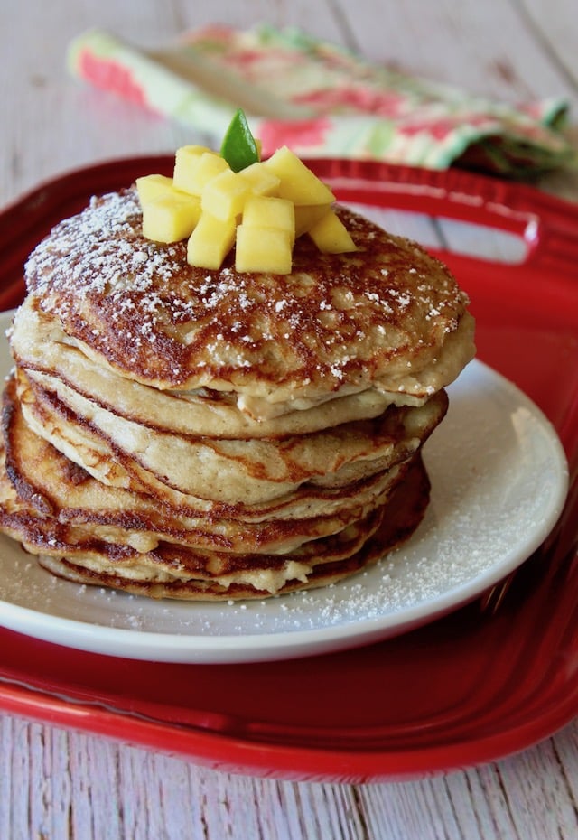 Tall stack of coconut flour pancakes on a whitle place iwth mango on top