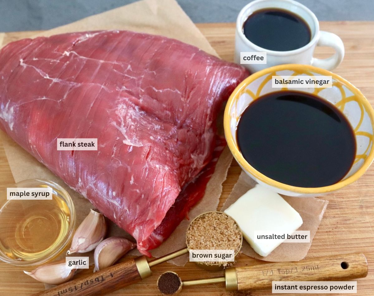 Raw flank steak, cup of coffee, tiny bowl of maple syrup, espresso, butter, brown sugar and balsamic sugar on a cutting board.