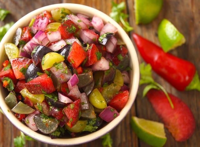 Grape-Plum Summer Salsa in a white rimmed bowl surrounded with lime and plum slices on wood sruface.