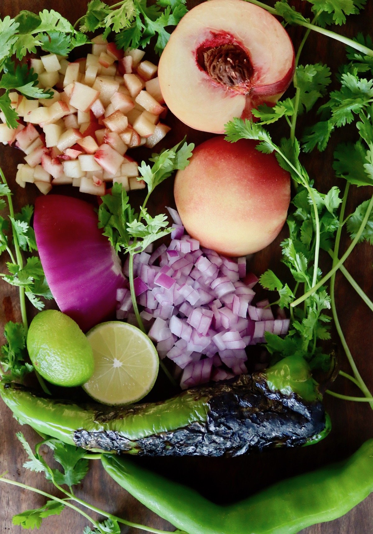 White peach, halves and chopped, fresh cilantro, chopped red onion, hatch chiles and halved lime on wood cutting board.