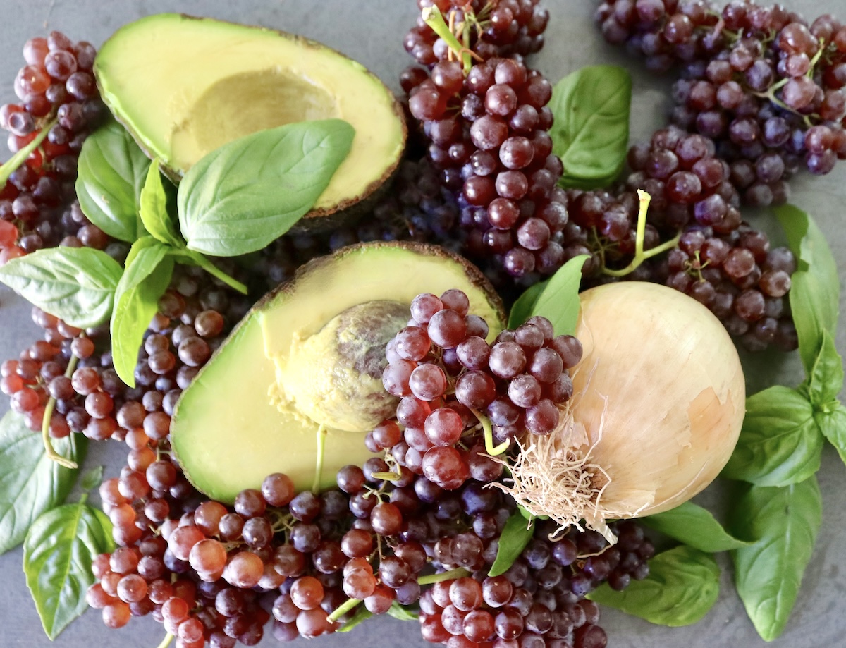 Ingredients for grape burschetta including an avocado sliced in half, onion, basil and champagne grapes in clusters.
