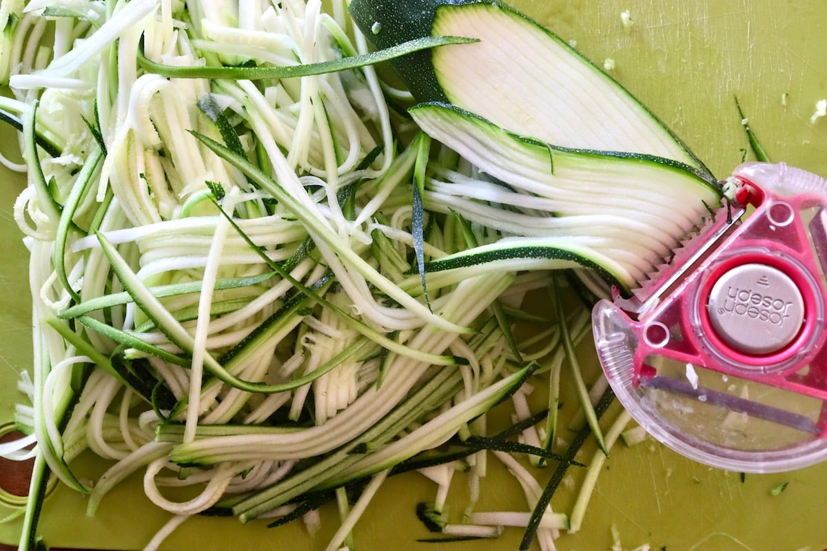 Spiralized zucchini noodles on a green plastic cutting sheet with a pink julienne peeler.