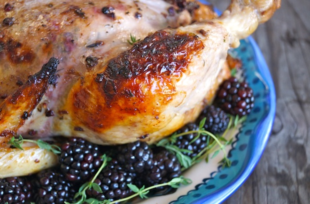 Blackberry-Chicken-Recipe-with-Blackberry-Butter) Close up of the side of a whole Roasted Chicken with Blackberry Butter and fresh blackberries and fresh thyme, on a blue-rimmed ceramic platter.