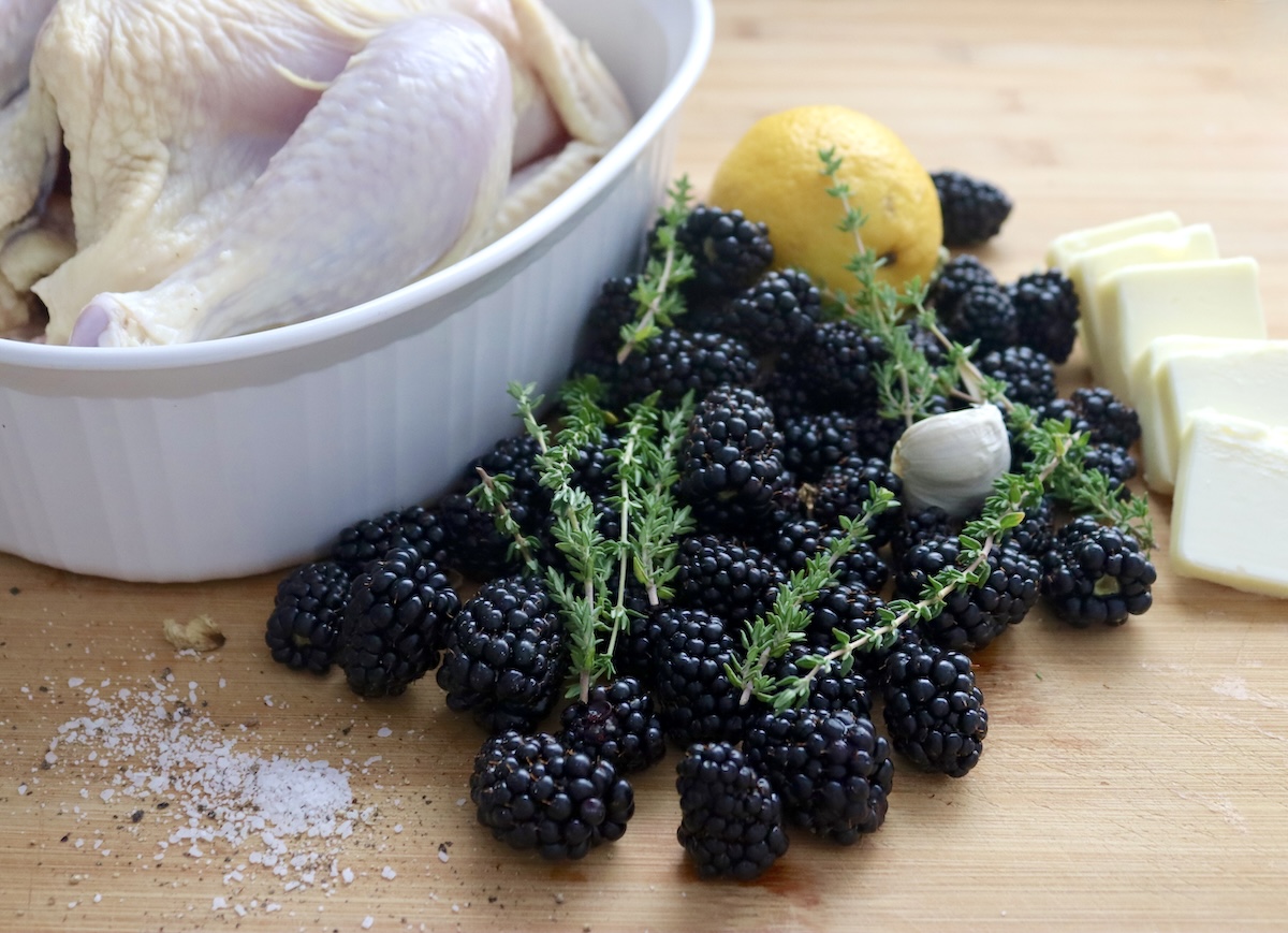 Blackberry-Chicken-Recipe-with-Blackberry-Butter Whole raw chicken in a white roasting dish with a pile of fresh blackberries, one garlic clove, thyme sprigs, a lemon and 6 pats of butter next to it.