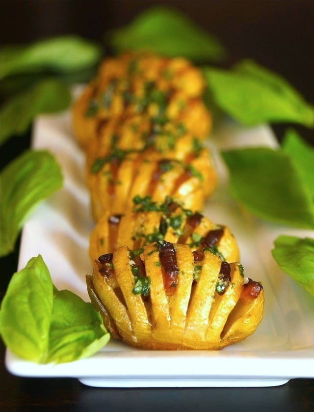 Several Bacon Dutch Yellow Hasselback Potatoes on a long, white narrow plate surrounded with fresh basil leaves