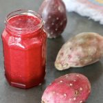 Jar of prickly pear juice (cactus pear juice) with 3 whole prickly pears next to it, on gray surface.