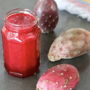 Jar of prickly pear juice (cactus pear juice) with 3 whole prickly pears next to it, on gray surface.
