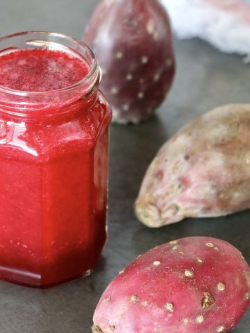 Jar of prickly pear juice (cactus pear juice) with 3 whole prickly pears next to it, on gray surface.