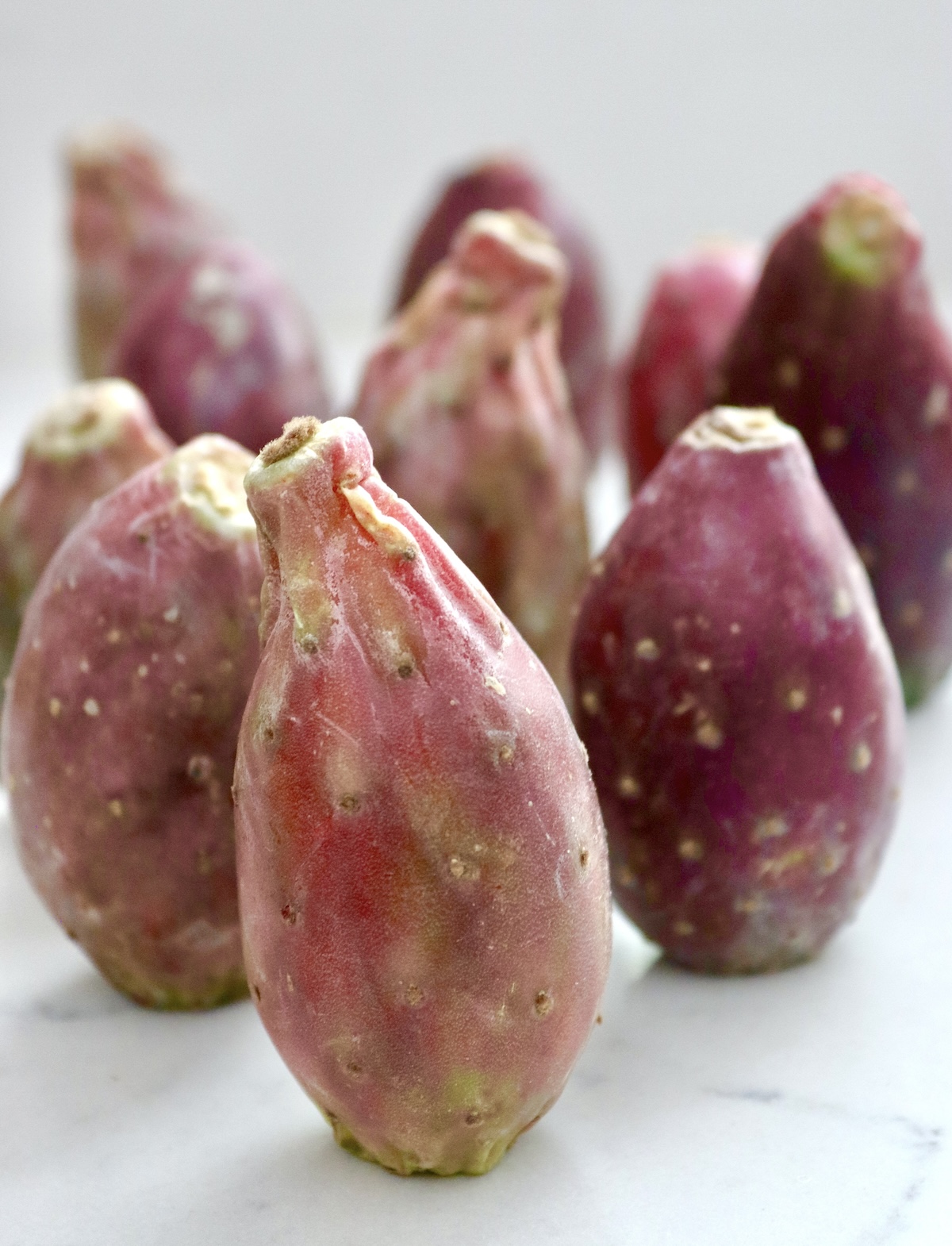 How-to-Cut-and-Eat-Cactus-Pears Several red prickly pears standing on their ends, on a white marble background.