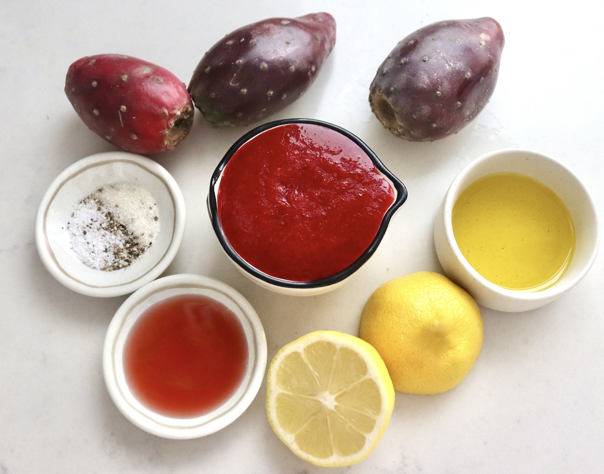 Prickly-Pear-Vinaigrette Ingredients for Prickly Pear Vinaigrette on a white marble surface, including, the prickly pear juice, lemon, red wine vinegar, extra virgin olive oil, salt, sugar, pepper and a few of the whole fruits in the background.