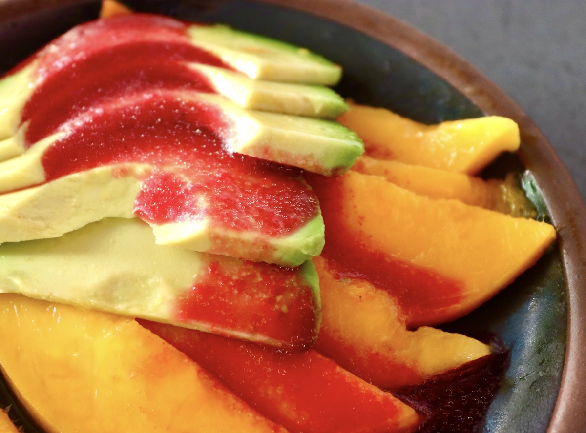 Prickly-Pear-Vinaigrette-with-Fruit Small black plate with sliced mango and avocado, with prickly pear vinaigrette drizzled over it.