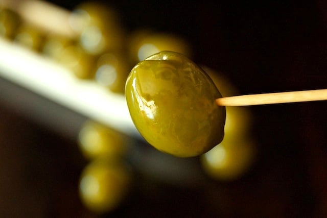 Warm Castelvetrano Olives in a narrow, white dish in the background of one of the green olives on a toothpick.