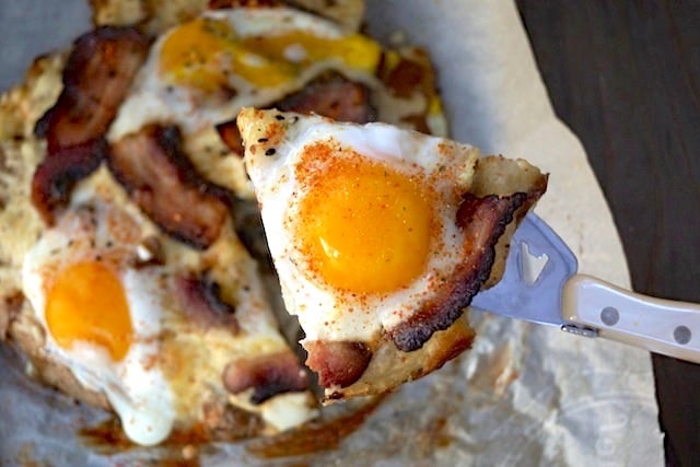 Slice of Smashed Potato Egg Pizza with Bacon being held up high above whole pizza